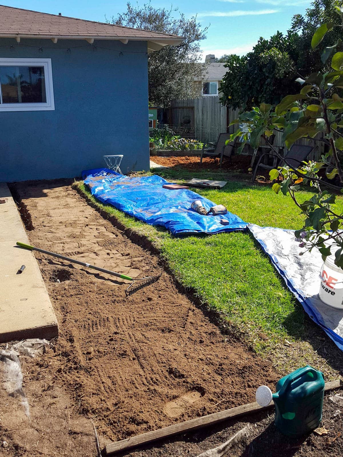 A section of grass has been removed down to bare earth around the edge of a concrete patio so raised beds can be placed on top of it to start a new garden. 