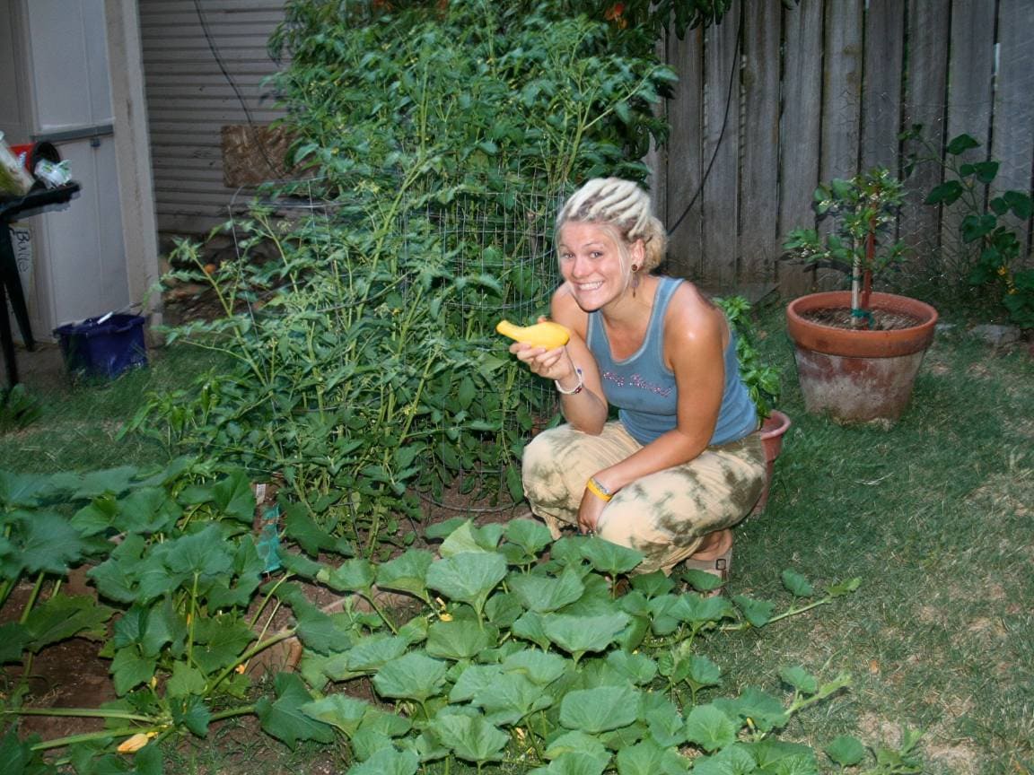 DeannaCat sitting amongst growing squash, melons, and tomatoes. She is holding a freshly harvest squash in her first garden. 