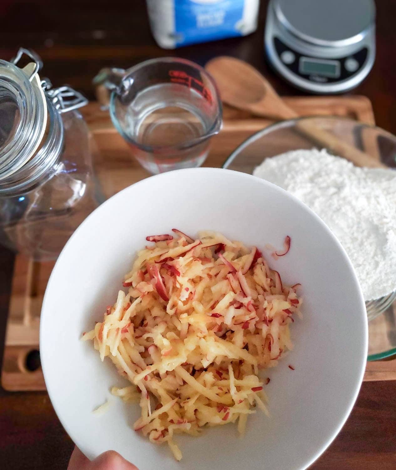 A white ceramic bowl full of grated fresh apple. A bowl of flour and a measuring cup of of water is beyond. 