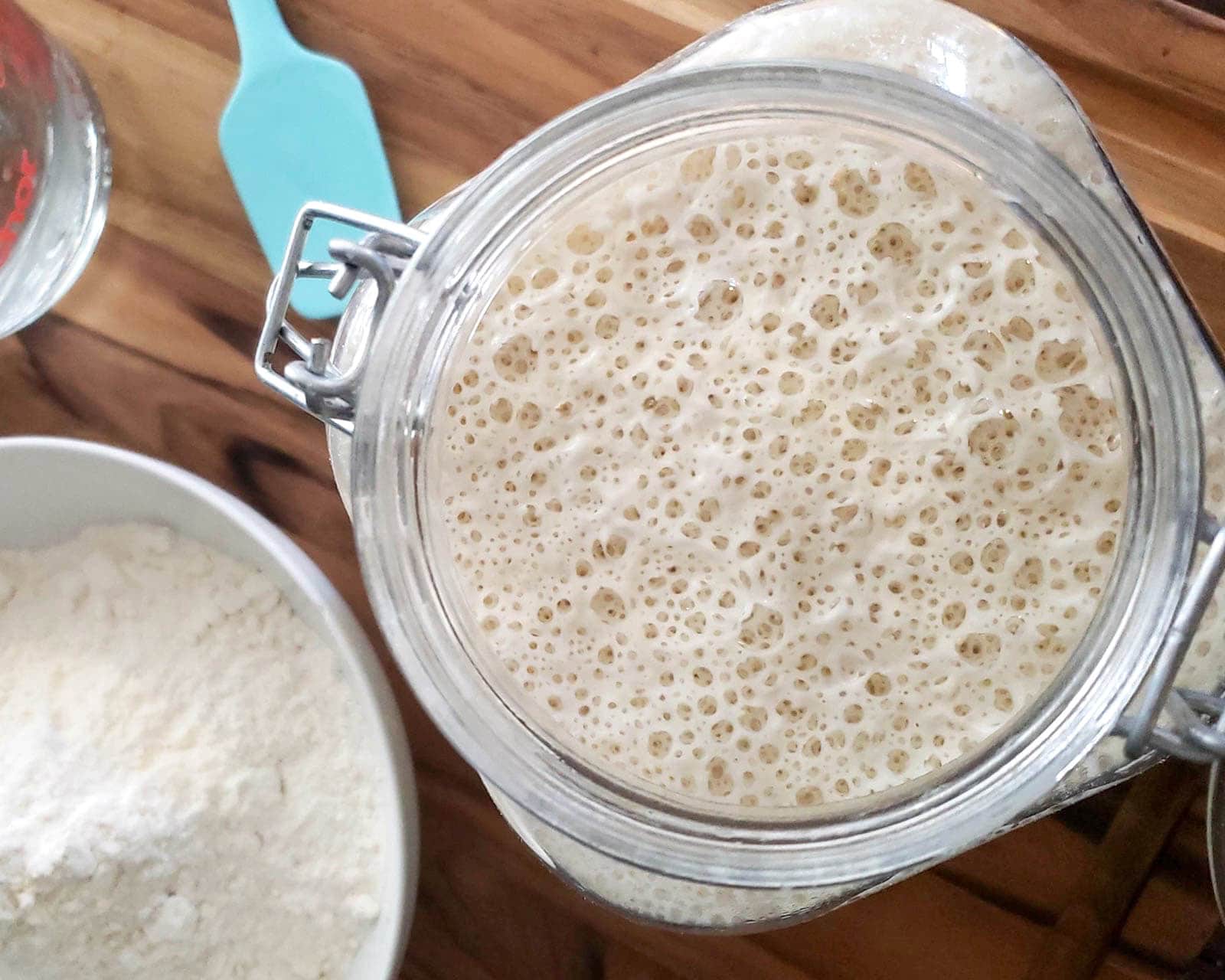 A large glass container full of a mixture of flour and water that is fluffy and risen towards the top of the jar. A bowl of flour and a blue silicone spatula sit nearby. 