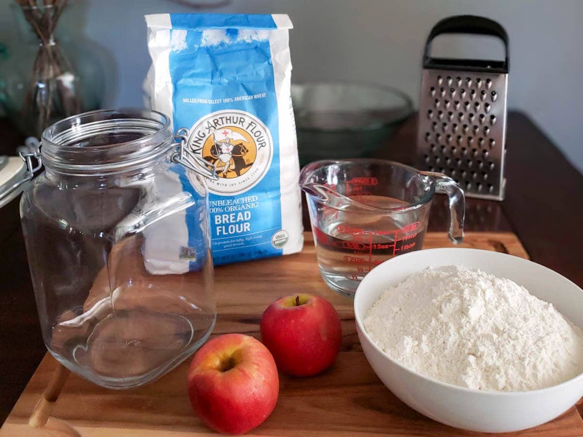 The supplies needed to make a sourdough starter sitting on a table: A large glass flip-top container, 2 small organic apples, a bowl of flour, filtered water in a measuring cup, a grater to be used on the apples, a scale to weigh the flour, and a large mixing bowl to combine it all. 