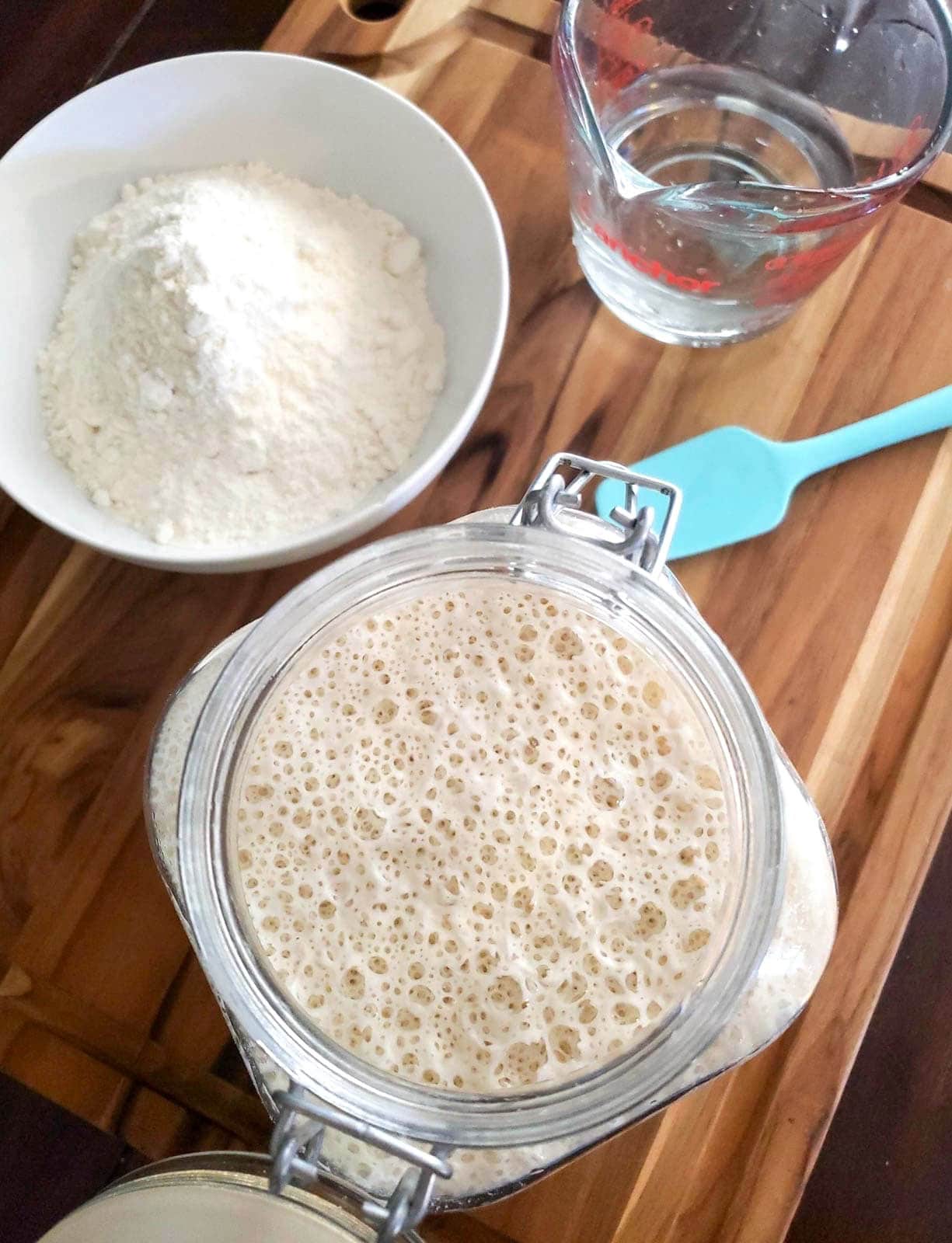 A large flip top glass jar is almost all the way full to the top with bubbly sourdough starter. A large ceramic bowl full of flower and a liquid glass measuring cup of water are nearby. 