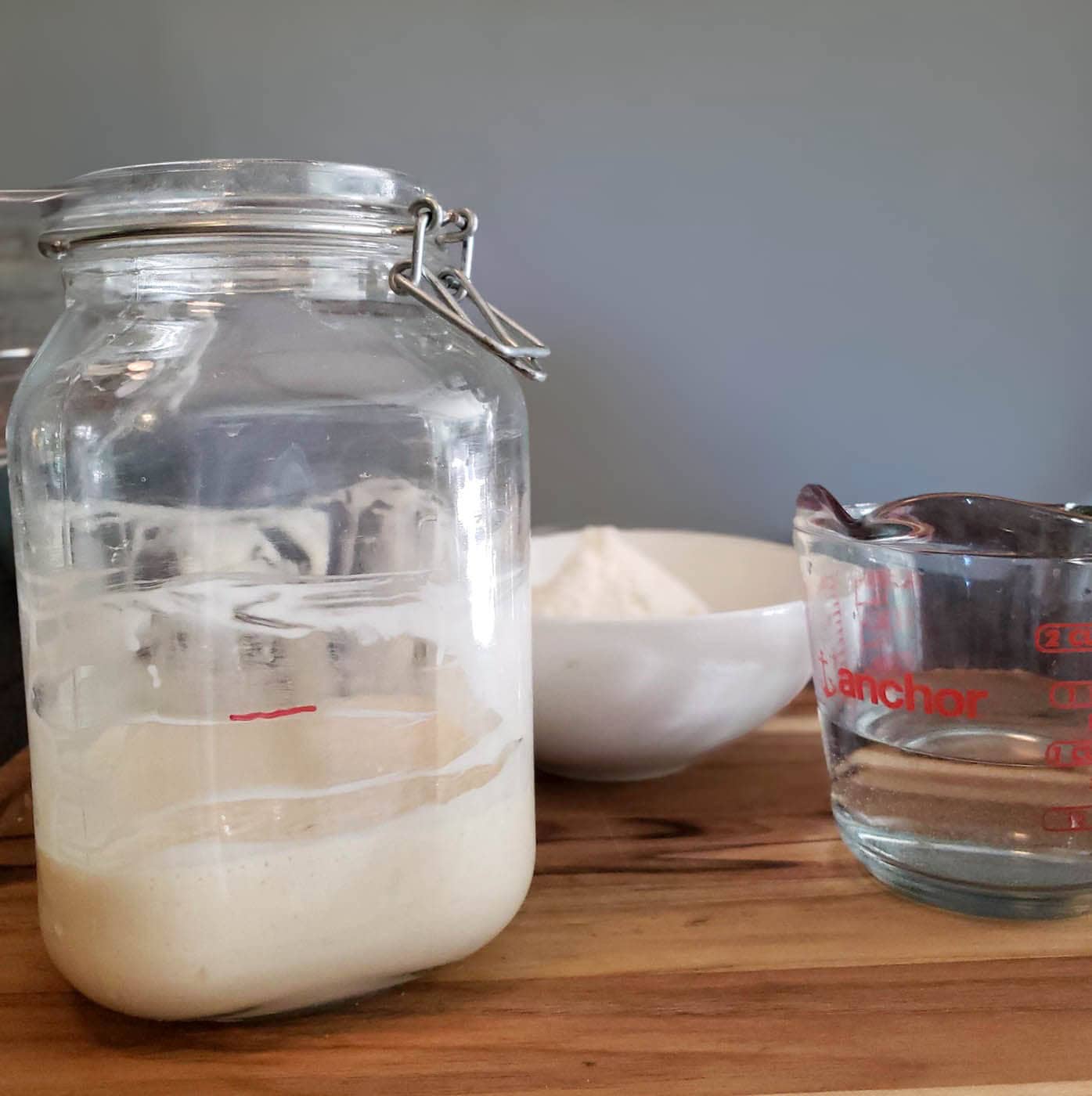 A glass container with a flip top lid contains a small amount of sourdough starter in the bottom of the container. A bowl of flour and a liquid glass measuring cup of water are nearby. 