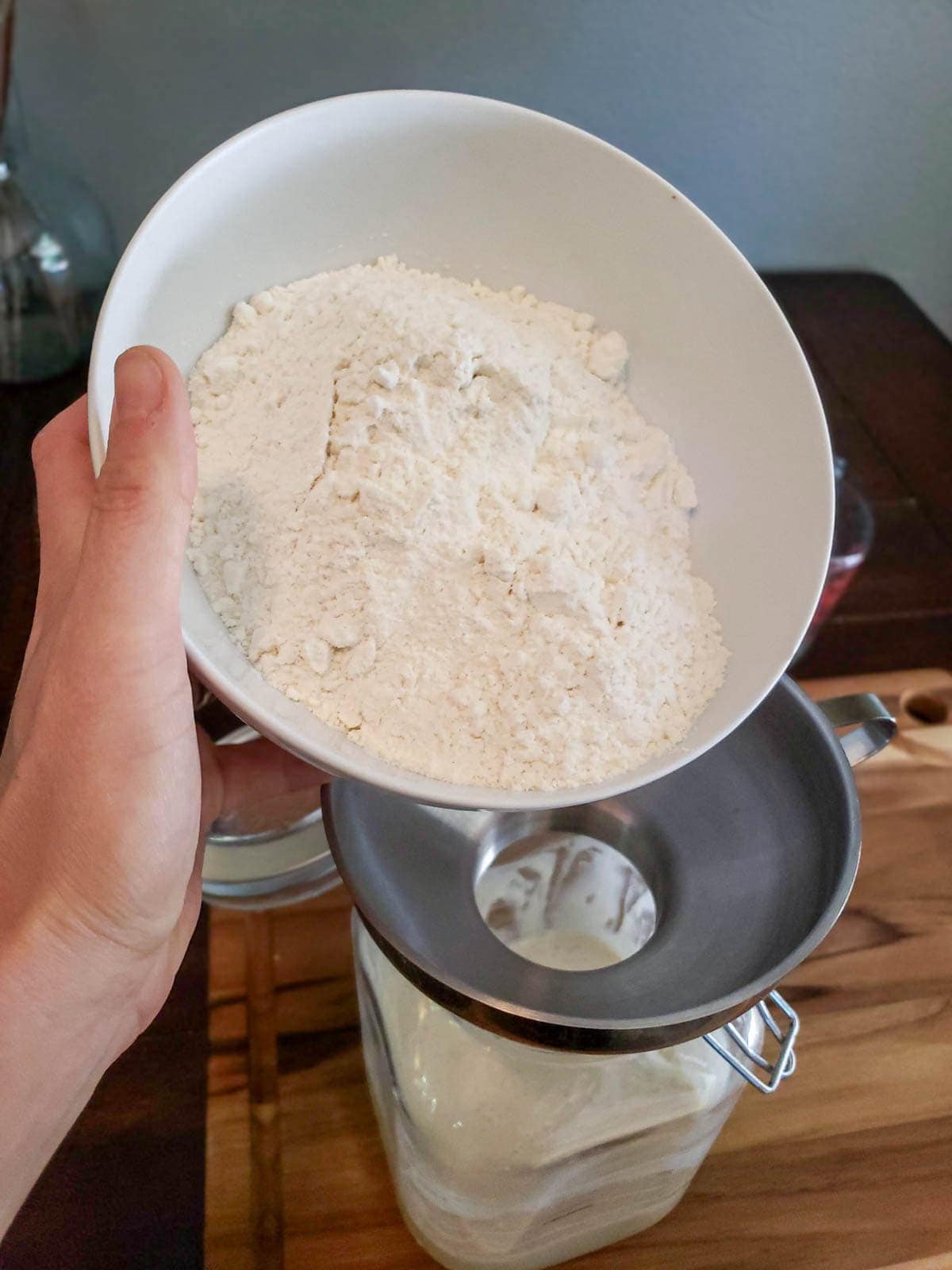 A ceramic bowl of flour is being poured through a metal funnel into a large glass container. 