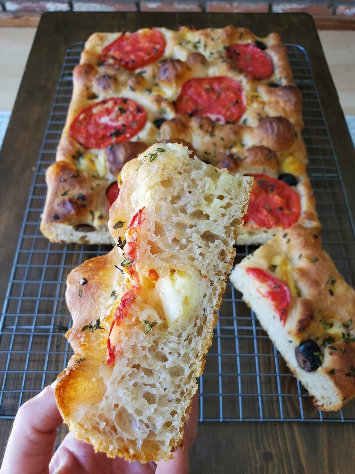 A hand is holding a slice of focaccia bread by the fingertips, it contains chunks of cheese, sliced tomatoes, black olives, and fresh herbs. Beyond is a wire cooling rack with the rest of the loaf of focaccia. 