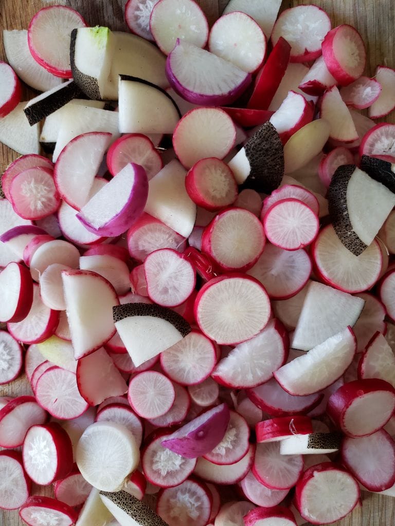 A cutting board piled high with slices  of homegrown radishes. Some are pink, red, and even with black skins!