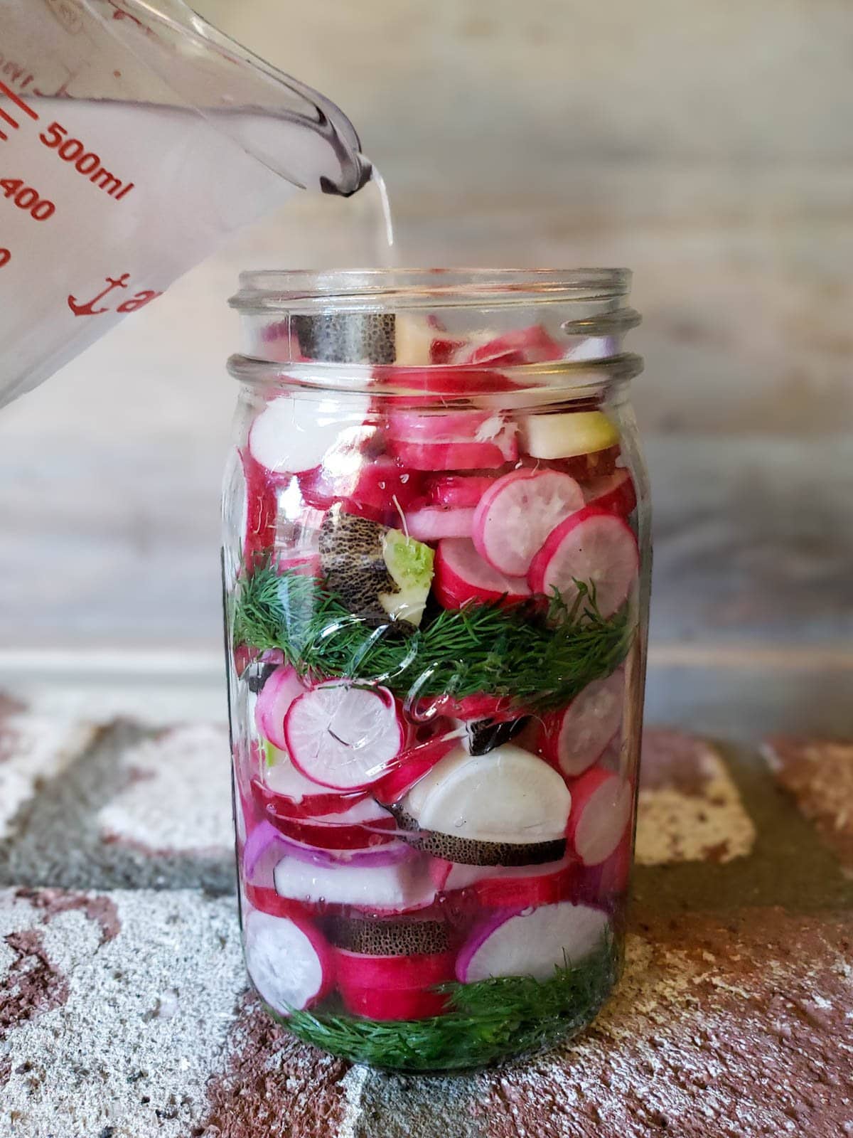 Salt water brine being poured from a glass measuring cup into the jar full of sliced radishes and dill