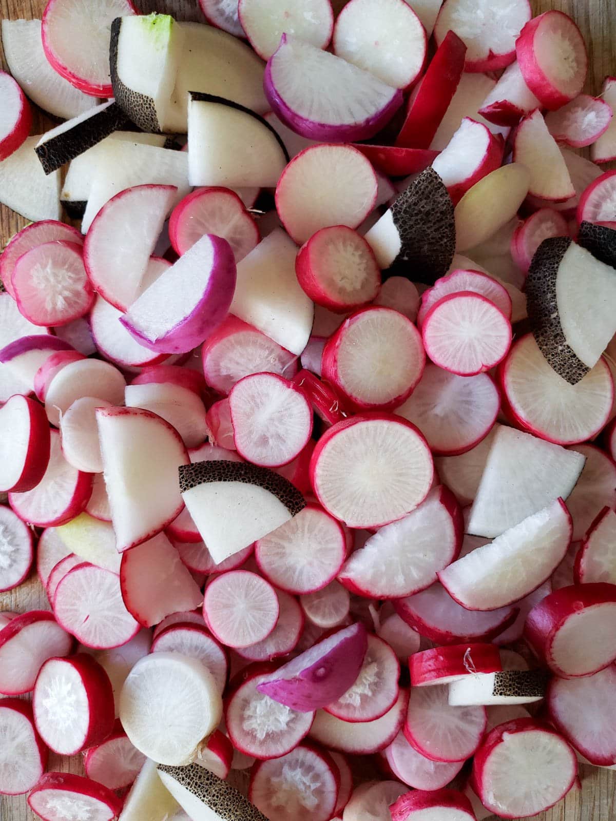 A cutting board piled high with fresh homegrown radishes cut into bite-size slices. Some are pink, red, and even with black skins!