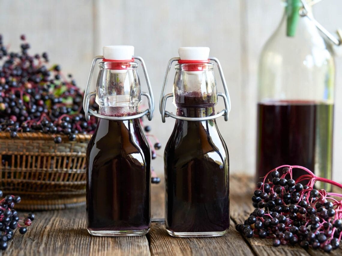 Two small glass flip-top bottles full of dark purple-black elderberry syrup, sitting on a wood table with baskets of fresh elderberries in the background and on the table.