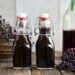 Two small glass flip-top bottles full of dark purple-black elderberry syrup, sitting on a wood table with baskets of fresh elderberries in the background and on the table.
