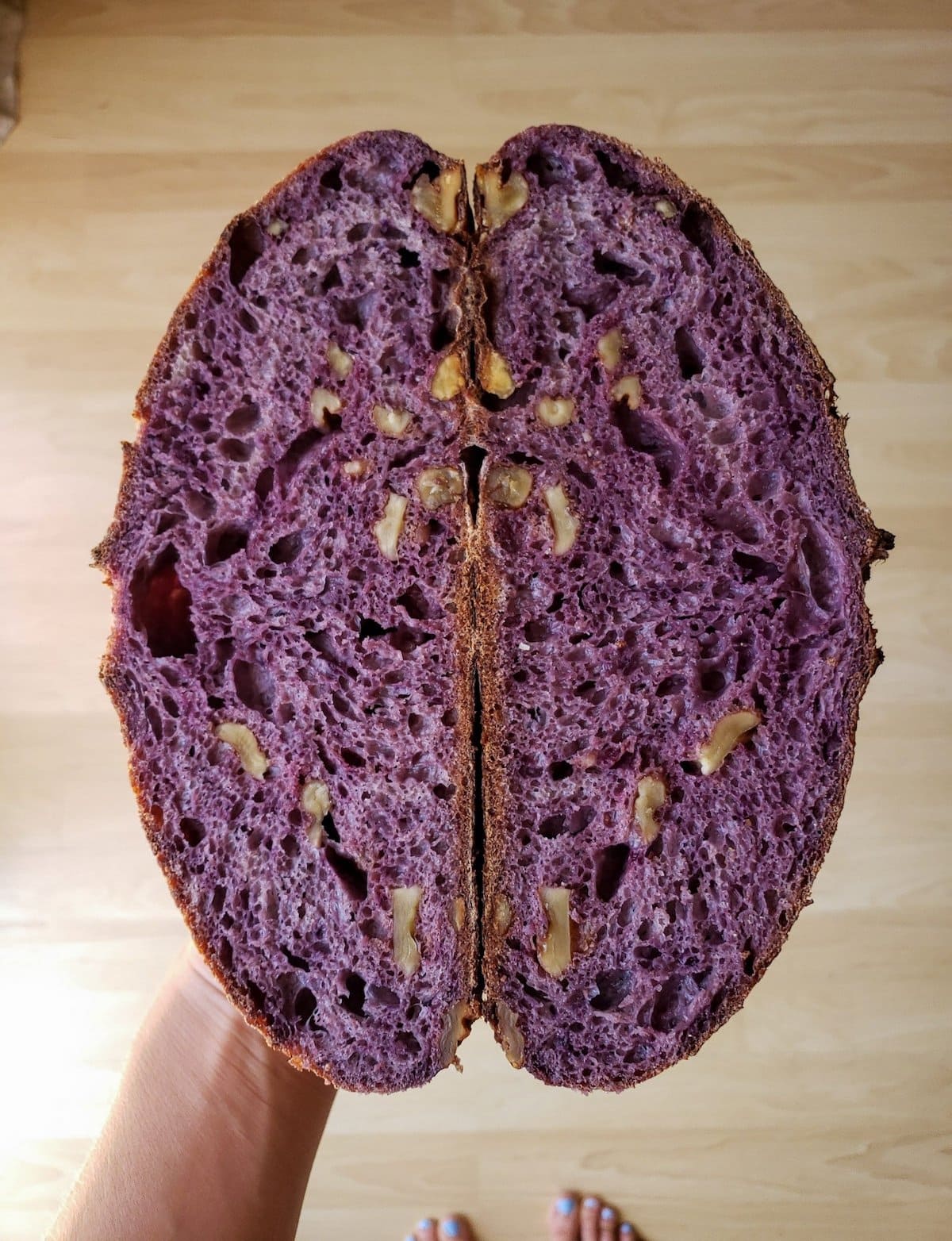 A loaf of purple sourdough bread with walnuts has been cut in half to show the inside of the bread which has been naturally colored using shredded purple carrots in the recipe. 