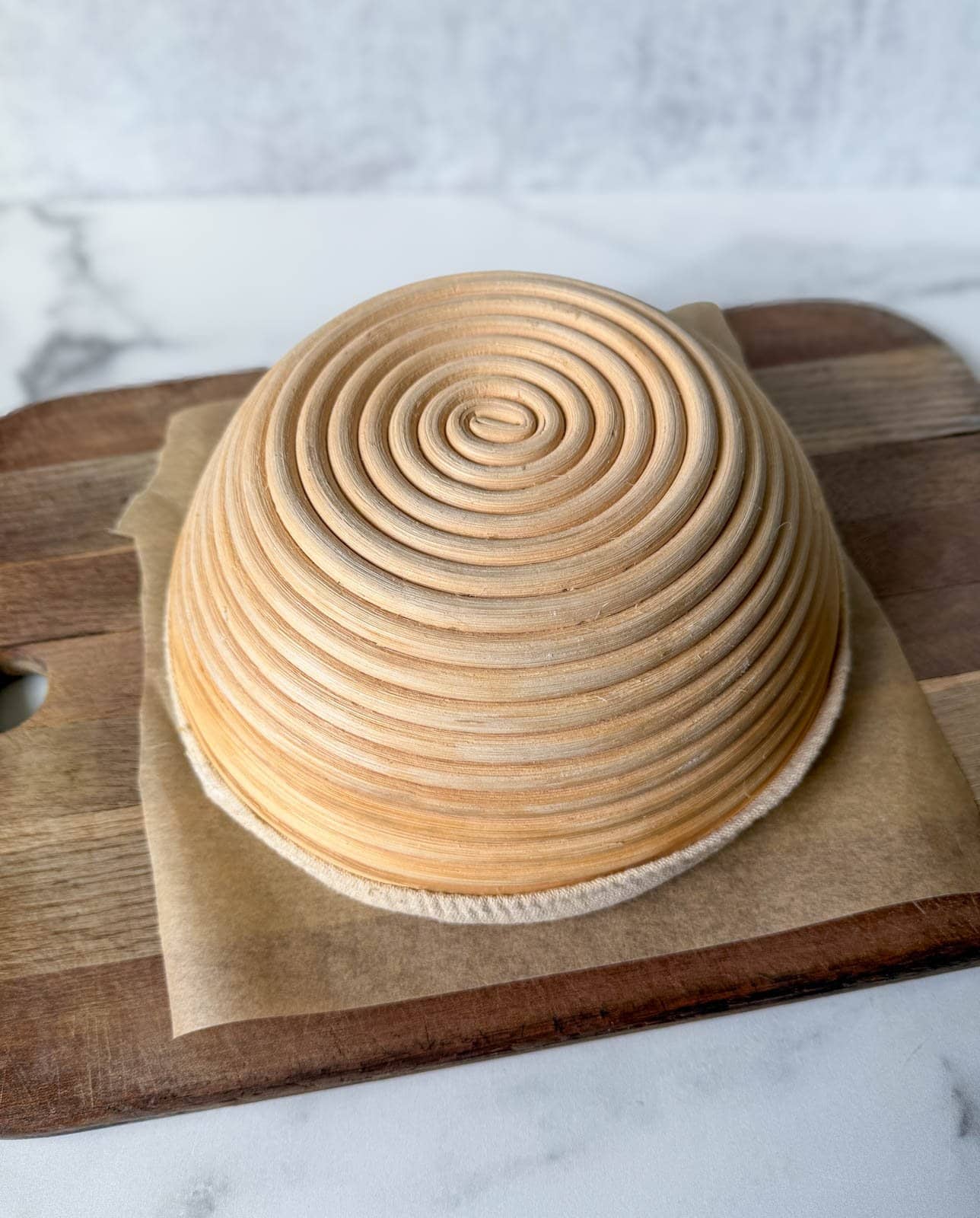 A banneton is sitting upside down on a cutting board with a piece of parchment paper in between the two.
