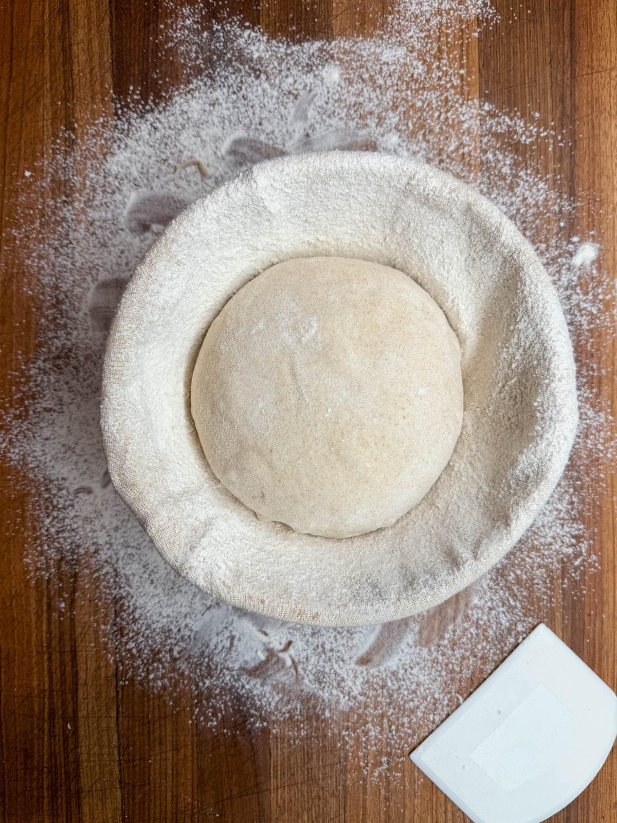 A dough ball is resting inside of a banneton which sits on a floured surface. 