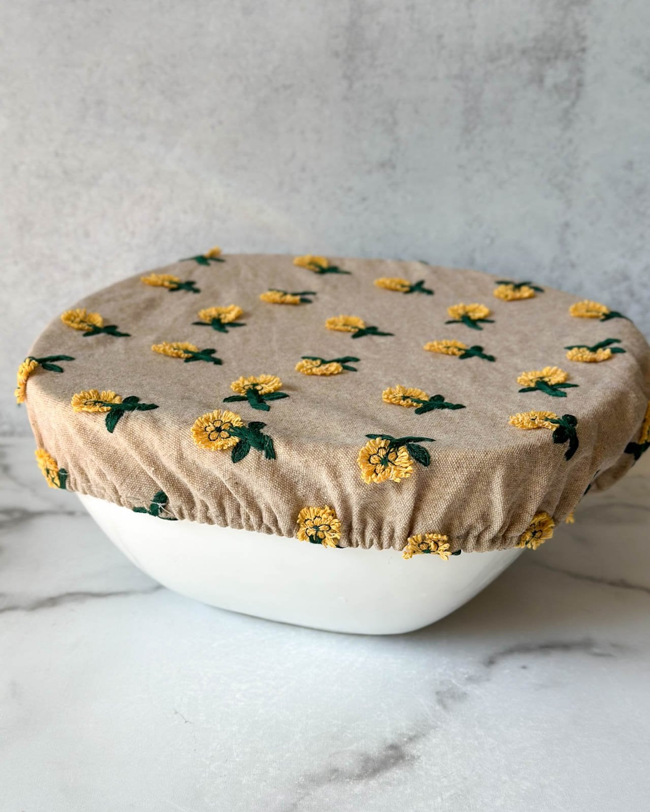 A large white ceramic bowl is covered with a damp cover with flowers stitched onto it.  