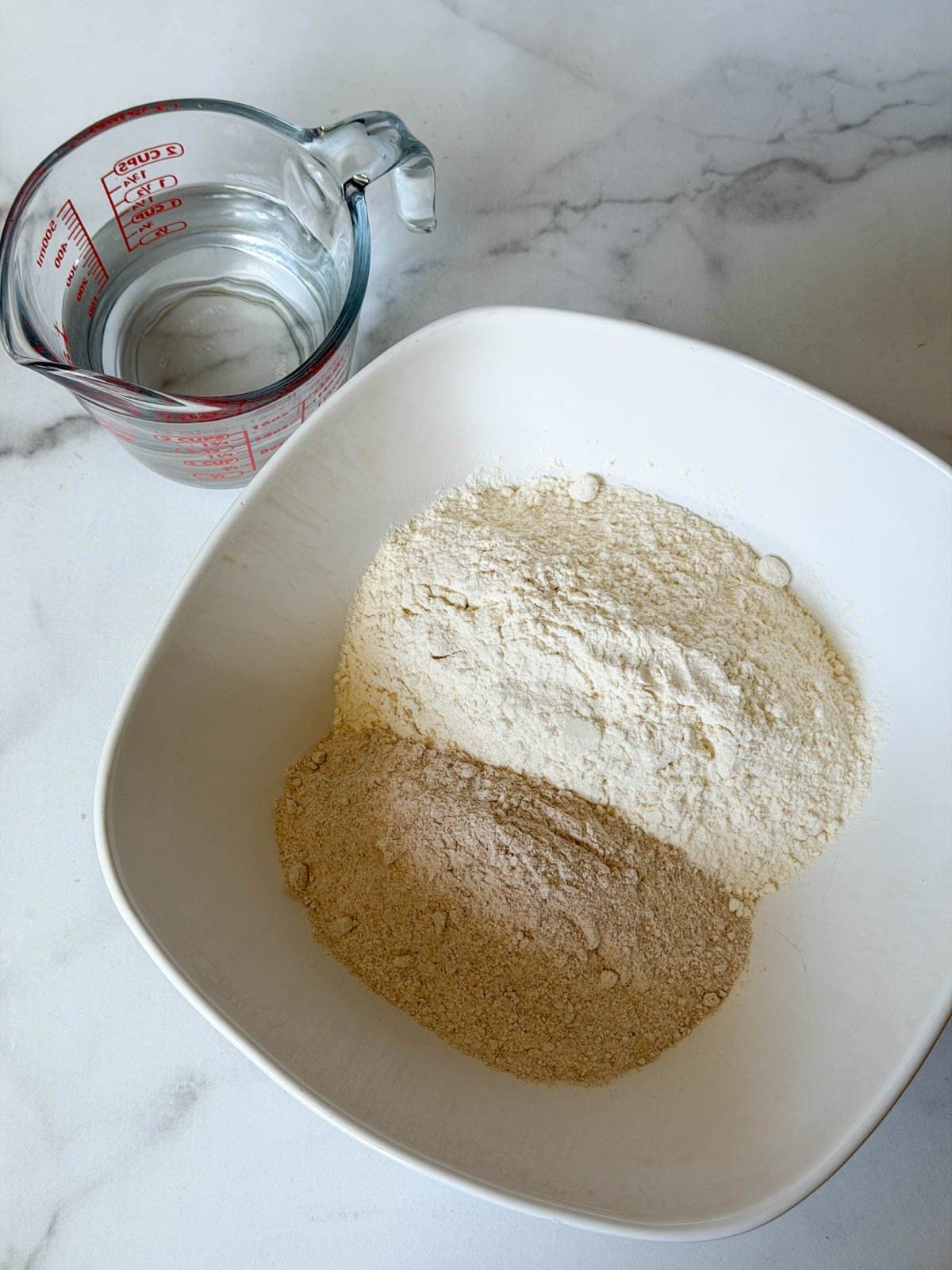 A large white bowl contains two types of flour while a liquid glass measuring cup of water is near. 