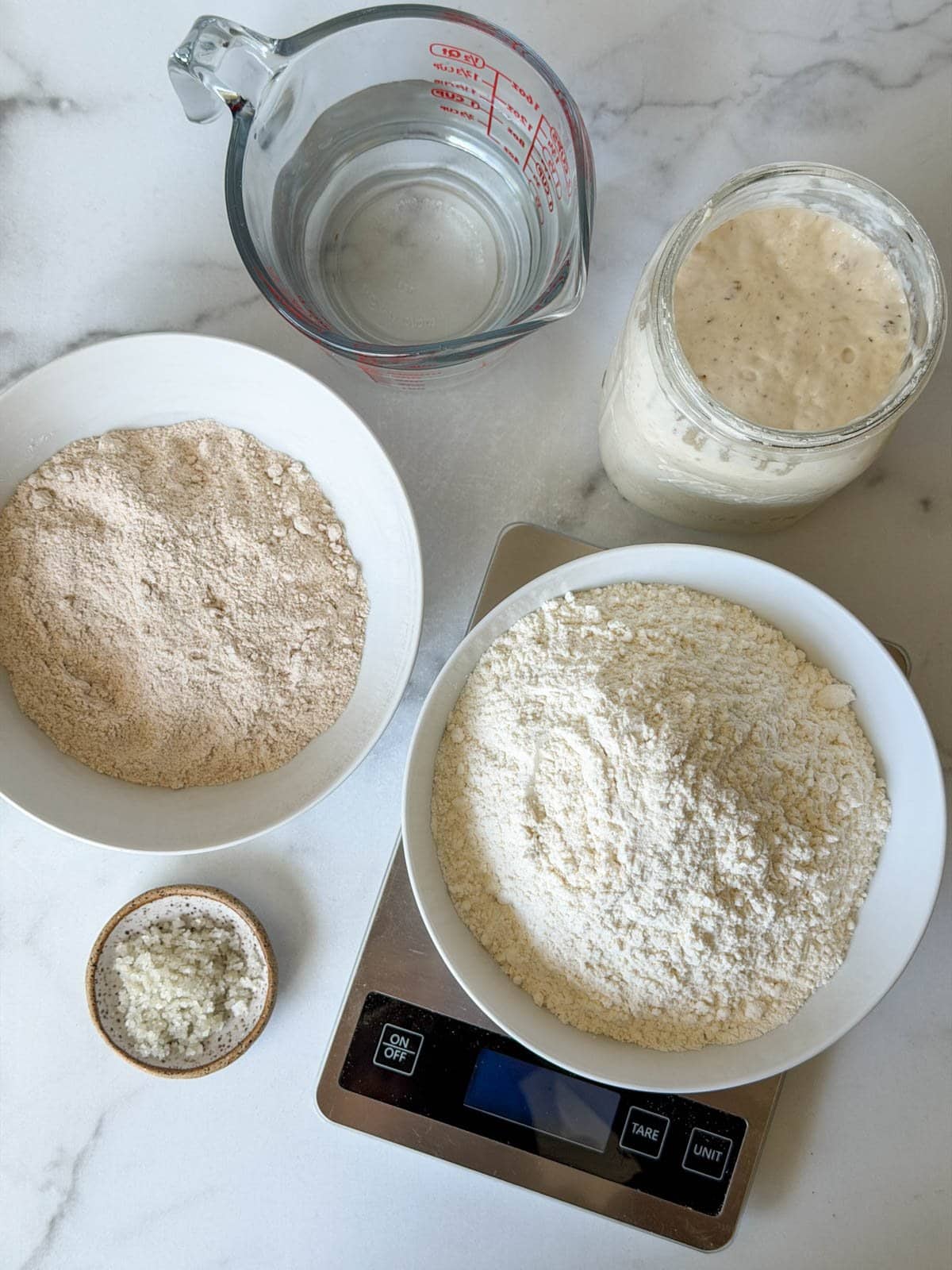 All of the ingredients to making sourdough bread, two bowls of flour, a jar of sourdough starter, a ramekin of salt, and a liquid measuring cup of water sit in a cluster together. 