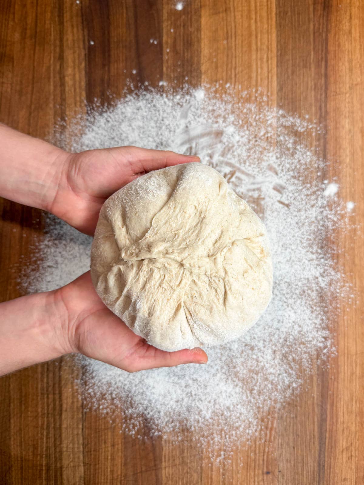 Two hands are holding a dough ball to show the seam in the dough above a floured surface. 
