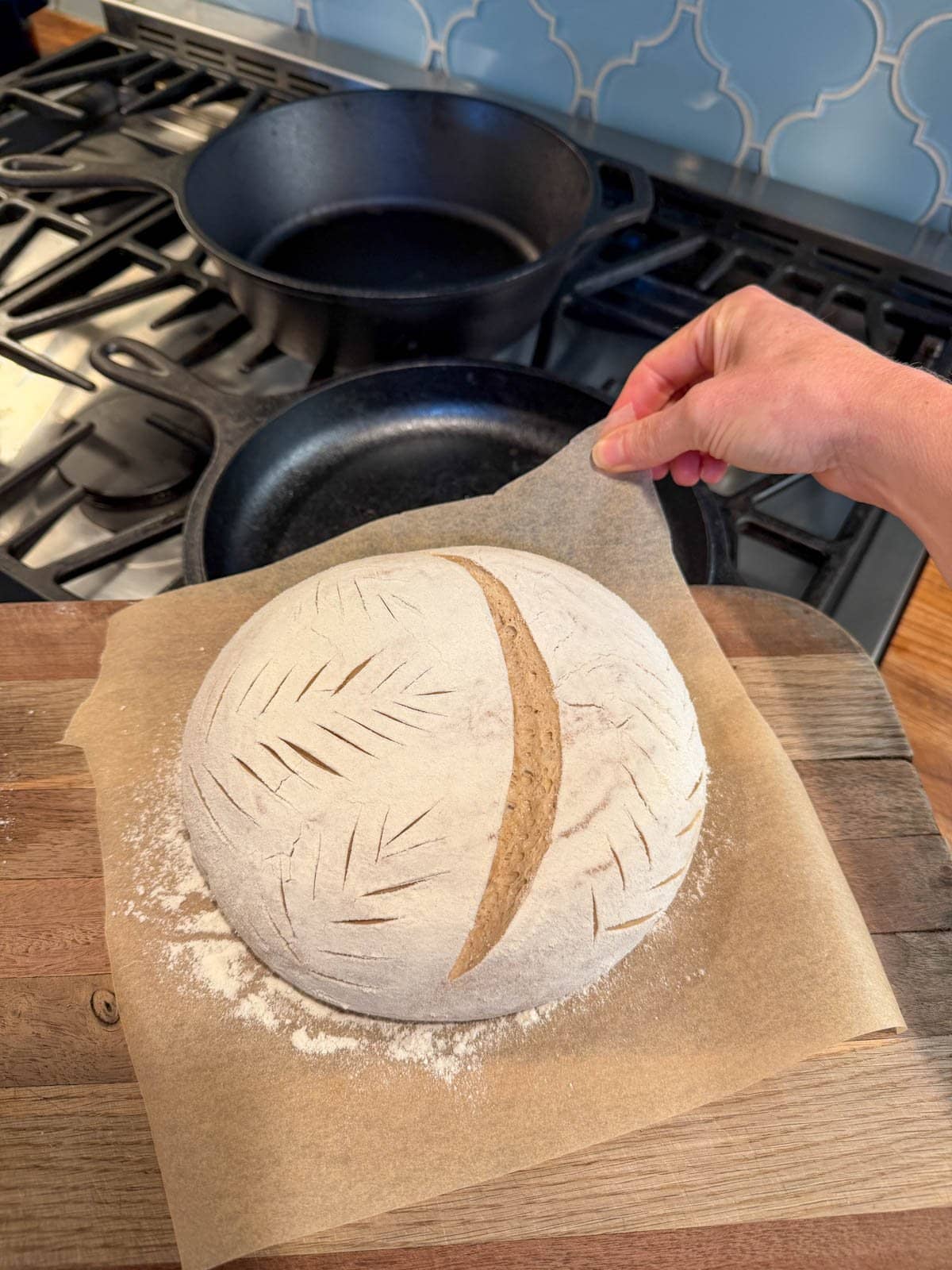 A loaf of uncooked sourdough bread, sitting on a cutting board. It is poised near a cast iron pan, ready to carefully be slid into it, and then into the oven. 