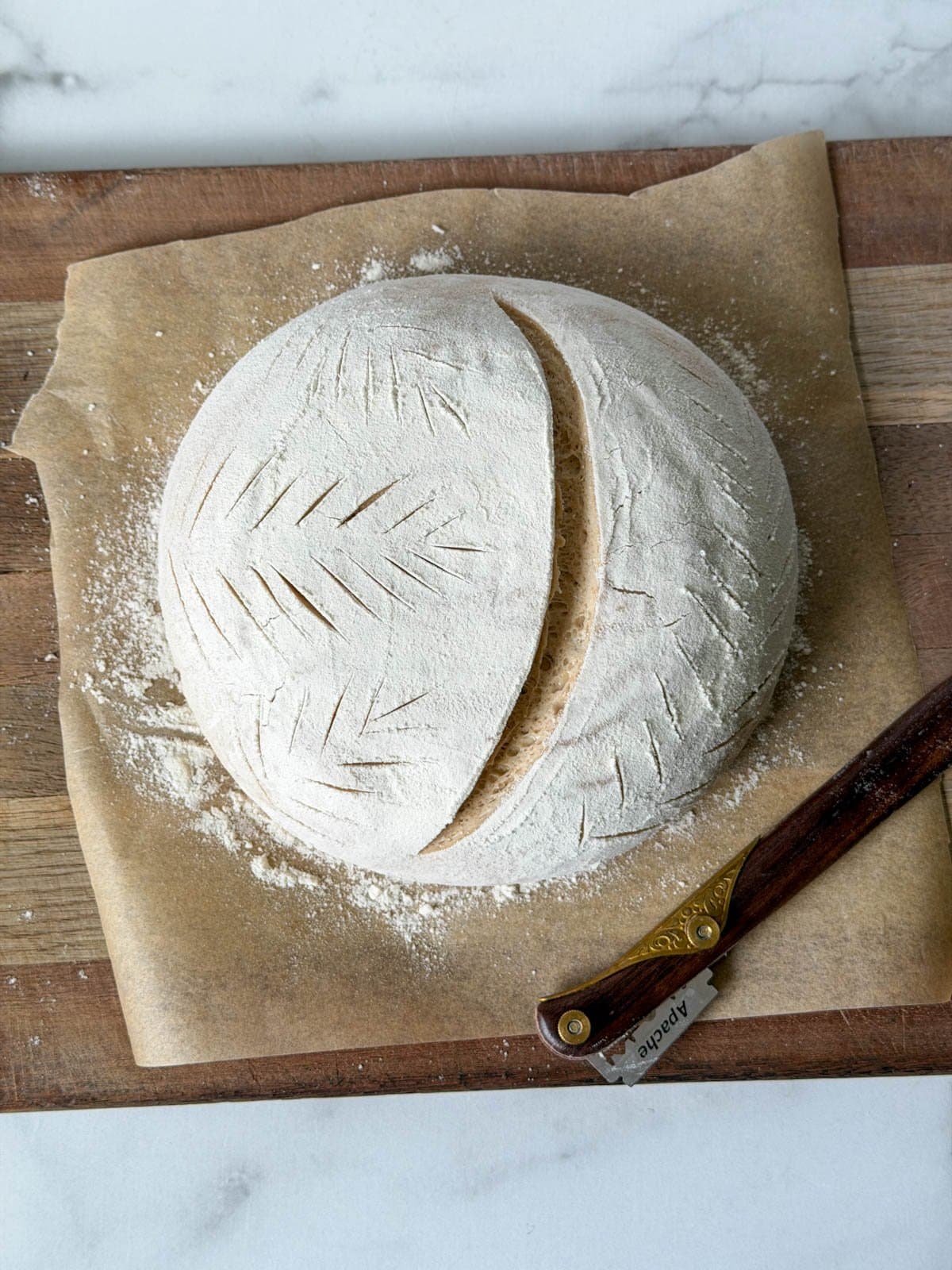 Dough in the shape of a boule is on a piece of parchment paper and has been scored in various places by a bread lame which is resting next to the dough. 