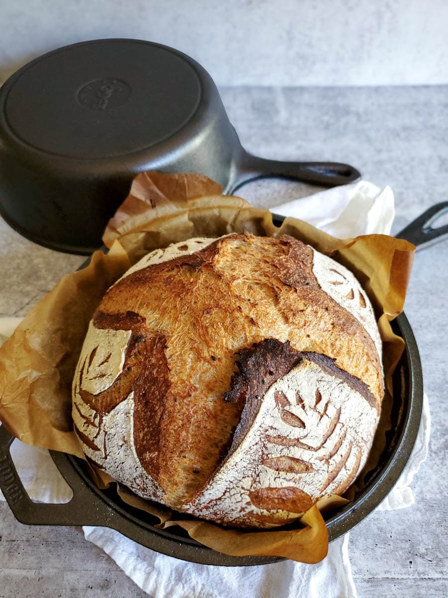 A loaf of freshly baked sourdough bread is shown still in its cast iron baking pan. The bread is light brown to darker brown in the more crispy parts. The strands of gluten are visible on the crust of the bread. 