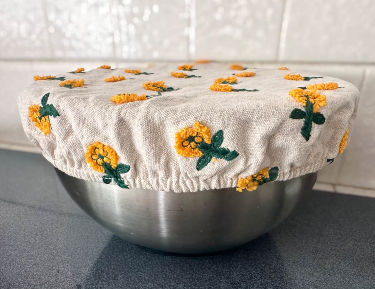 A large stainless steel bowl is partially covered with a cotton bowl cover that has flowers sewn into the fabric. 