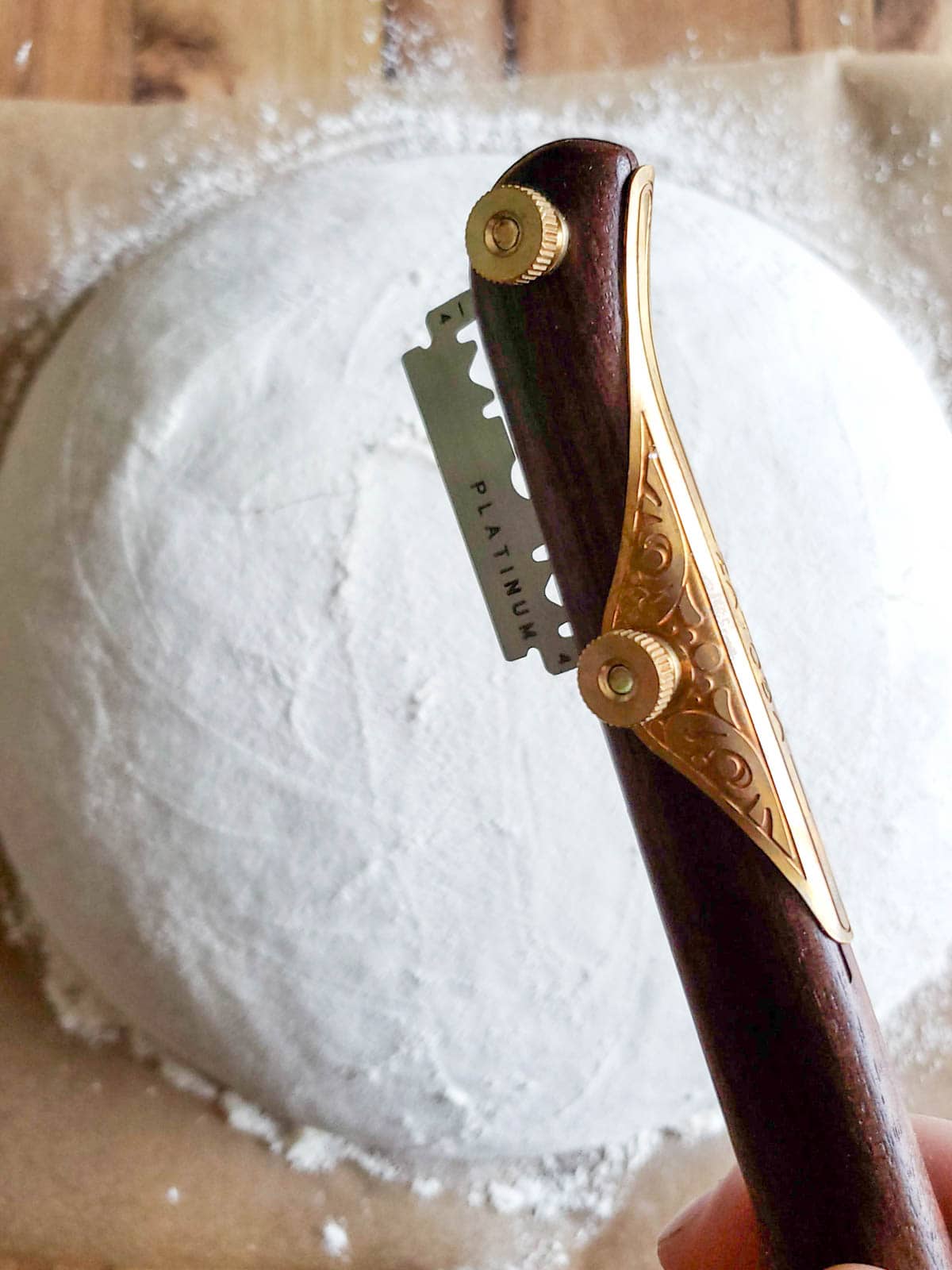 A wood handled bread lame is positioned above a loaf of raw sourdough bread sitting below. 