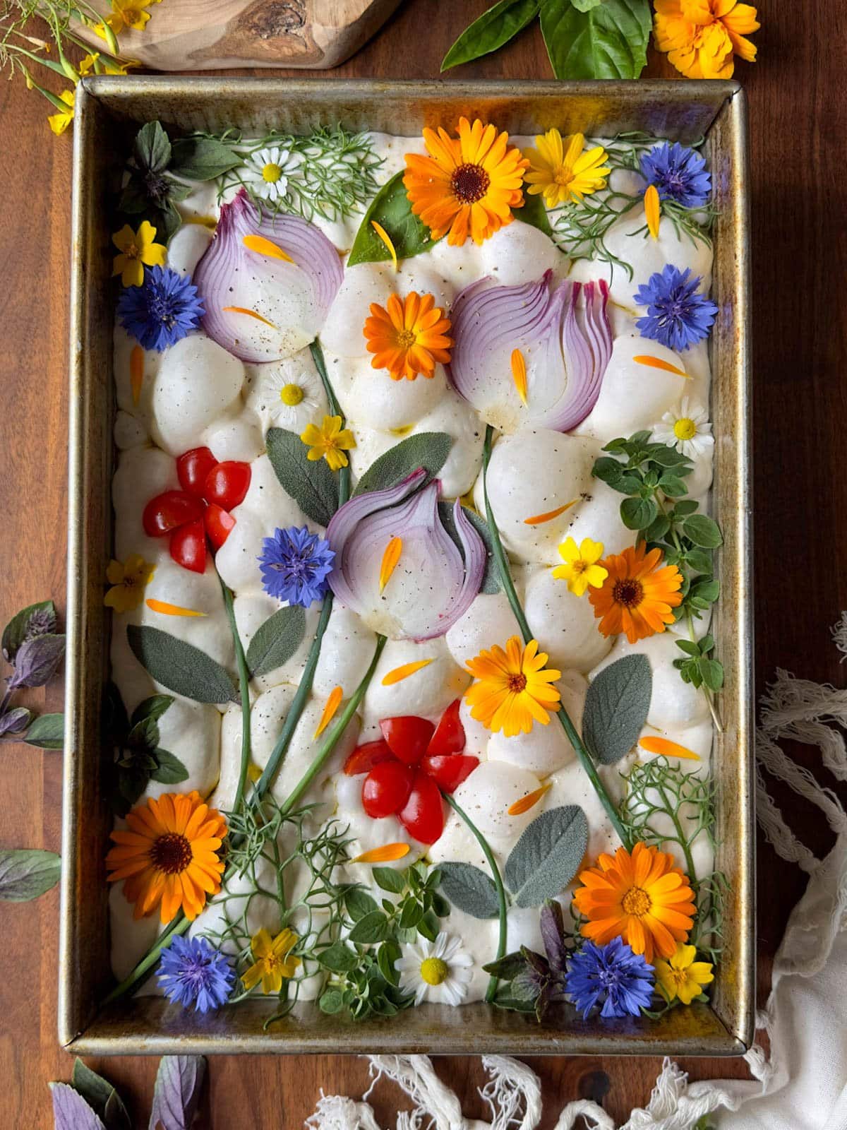 A cake pan contains a raw sourdough focaccia loaf with various veggies and herbs placed on top to create a nature theme with herbs used as stems with onions slices and tomatoes as flowers. 