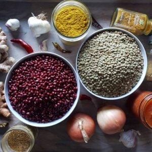 A photo of all the ingredients to make madras curry lentils. Two white bowls are full of red adzuki beans and green lentils, with onions, chillis, garlic, turmeric, ginger, and spices laying around the sides of the bowls
