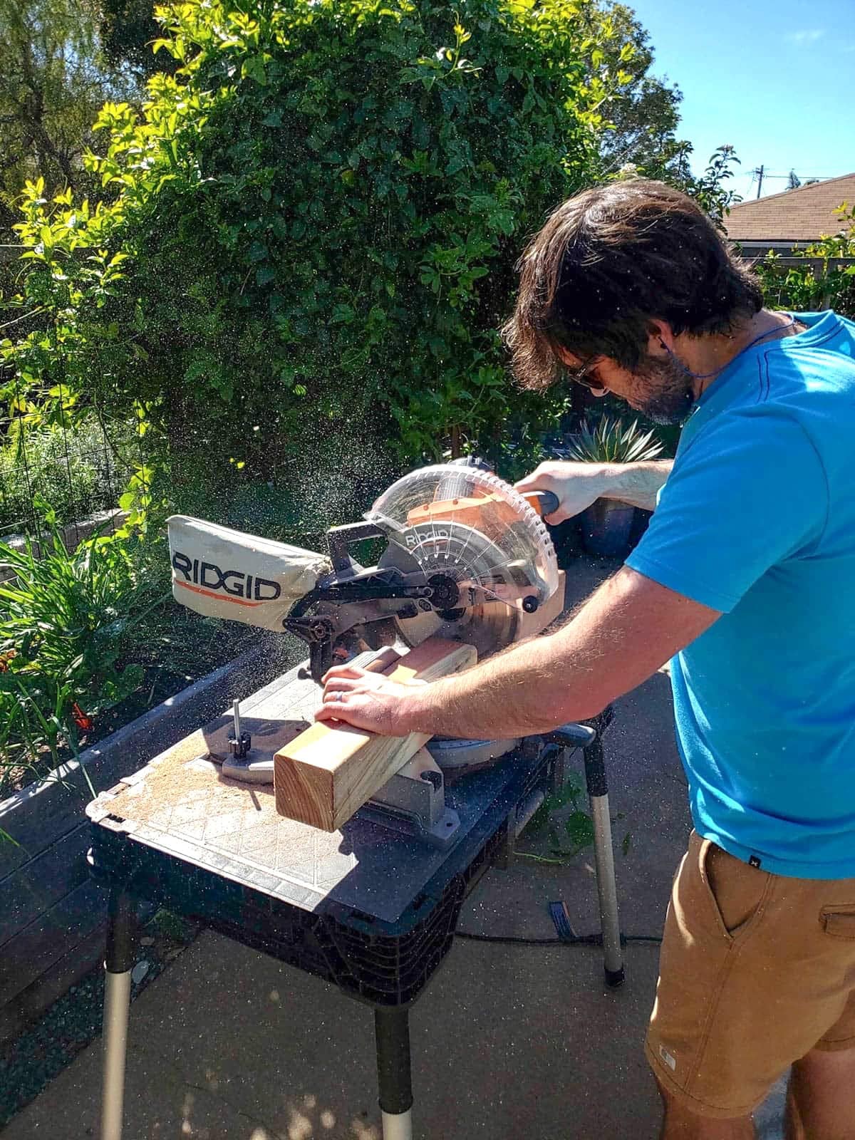 A man using a miter saw on a outdoor workbench cutting redwood boards for the raised garden bed