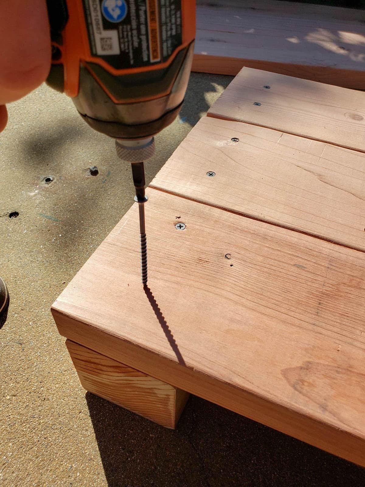 A drill being used to fasten screws to the side of the 2x6 boards, into the 4x4 corner pieces.