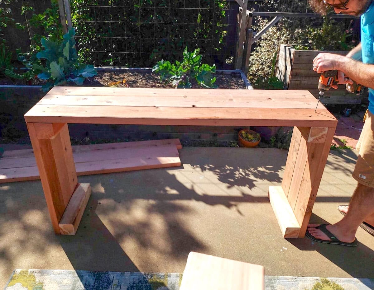 A man is using screws and a drill to attach longer boards to the short side that is standing on its end.