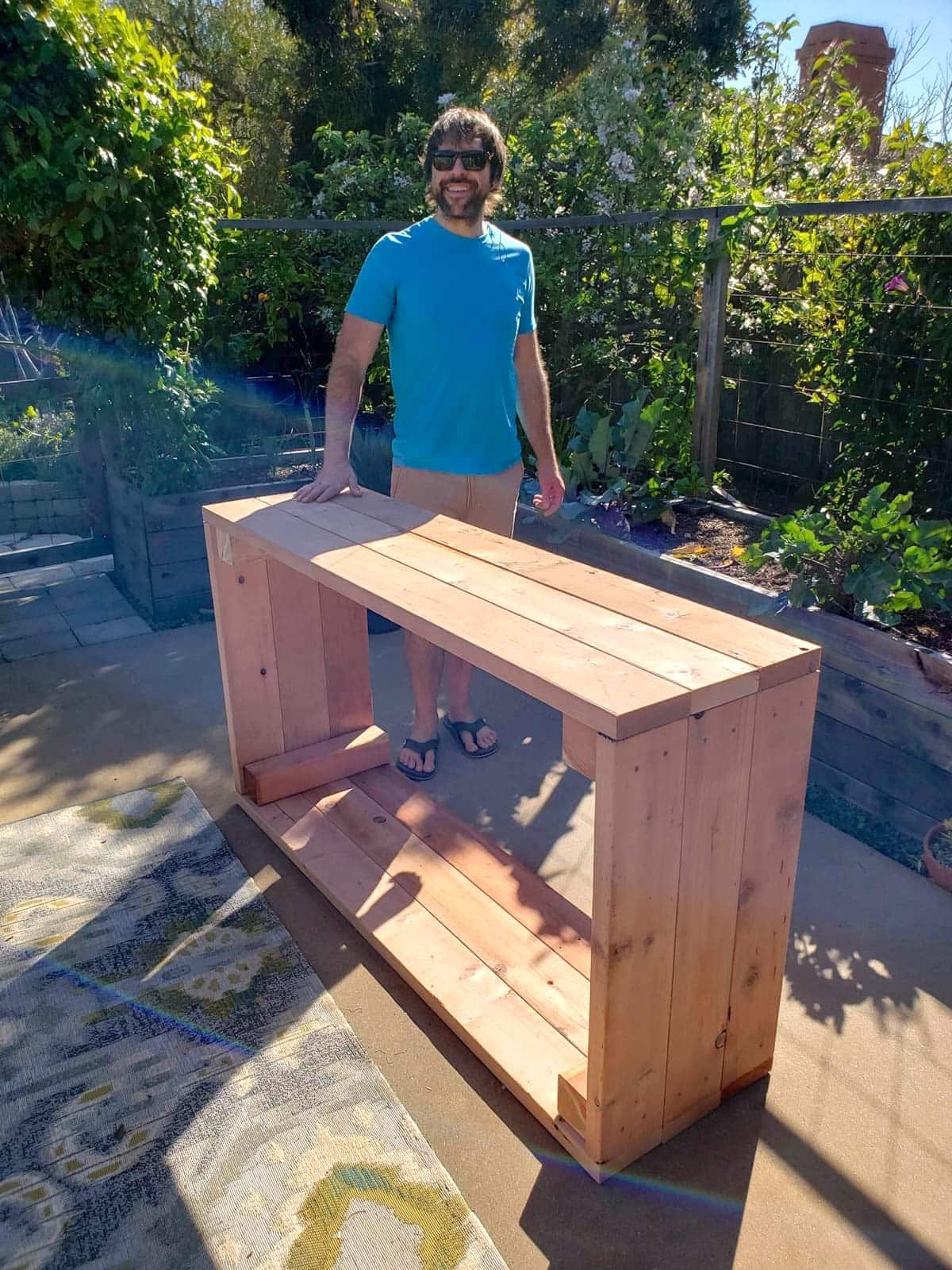 Aaron standing next to the just-built raised bed, still on the patio and on its side. He is smiling, wearing sunglasses, has dark brown hair, a short beard, and medium build. The patio garden and other raised beds (in use) are in the background.