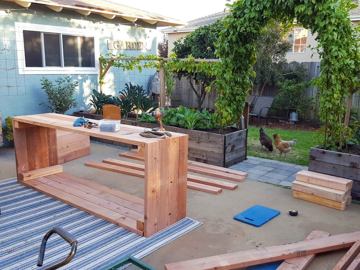 A backyard concrete patio contains cut lumber and one finished raised garden bed. Older garden beds surround the patio.