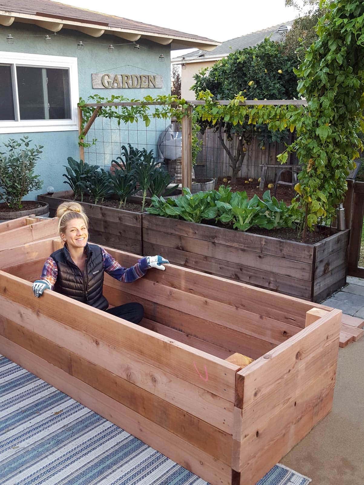 a young blonde woman sitting inside a large, deep, empty wooden raised garden bed frame sitting on a concrete patio, just built and not yet filled with soil