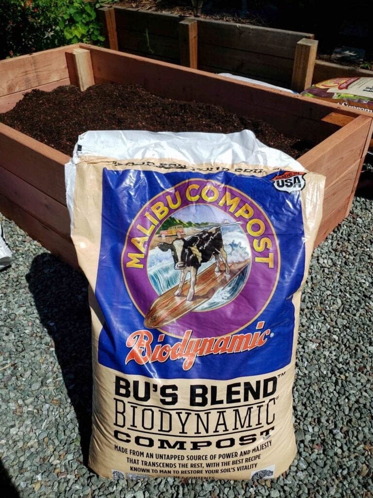 A photo of a bag of compost, by Malibu Compost. It is sitting on the gravel, leaning up against a half-filled raised bed behind it.