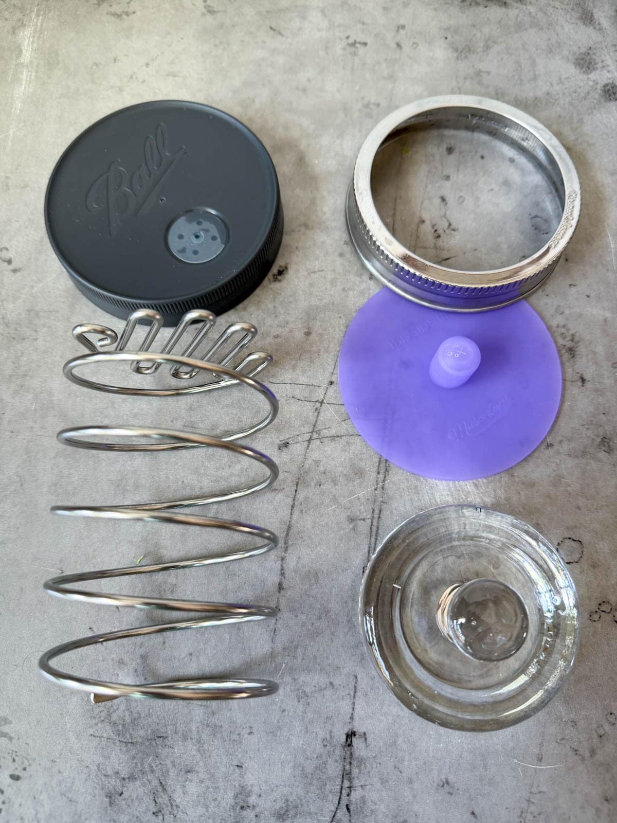 Two types of fermentation lids, one is a metal spring with a lid that contains holes for the release of gasses. The other is a glass weight with a pickle pipe top where the gasses will escape, along with a metal ring to secure it all in place to a jar.
