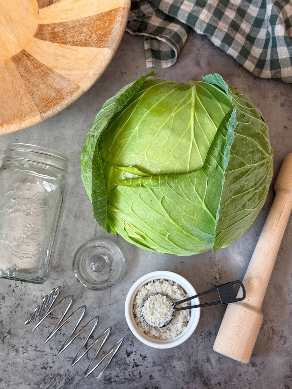 A large green head of cabbage sits amongst a ramekin of sea salt, a kraut pounder, a quart mason jar, a glass ferment weight, and a metal fermentation spring.