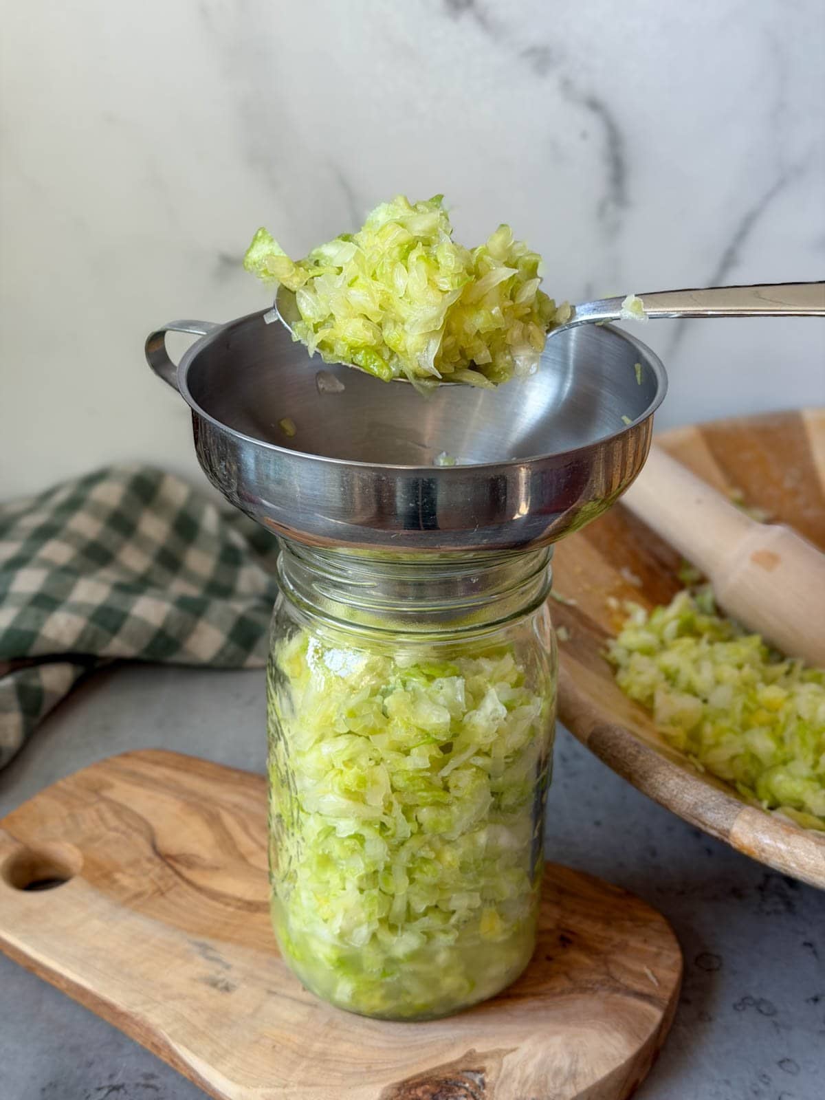 A large spoon is filling a quart jar with the assistance of a canning funnel full of salted cabbage.
