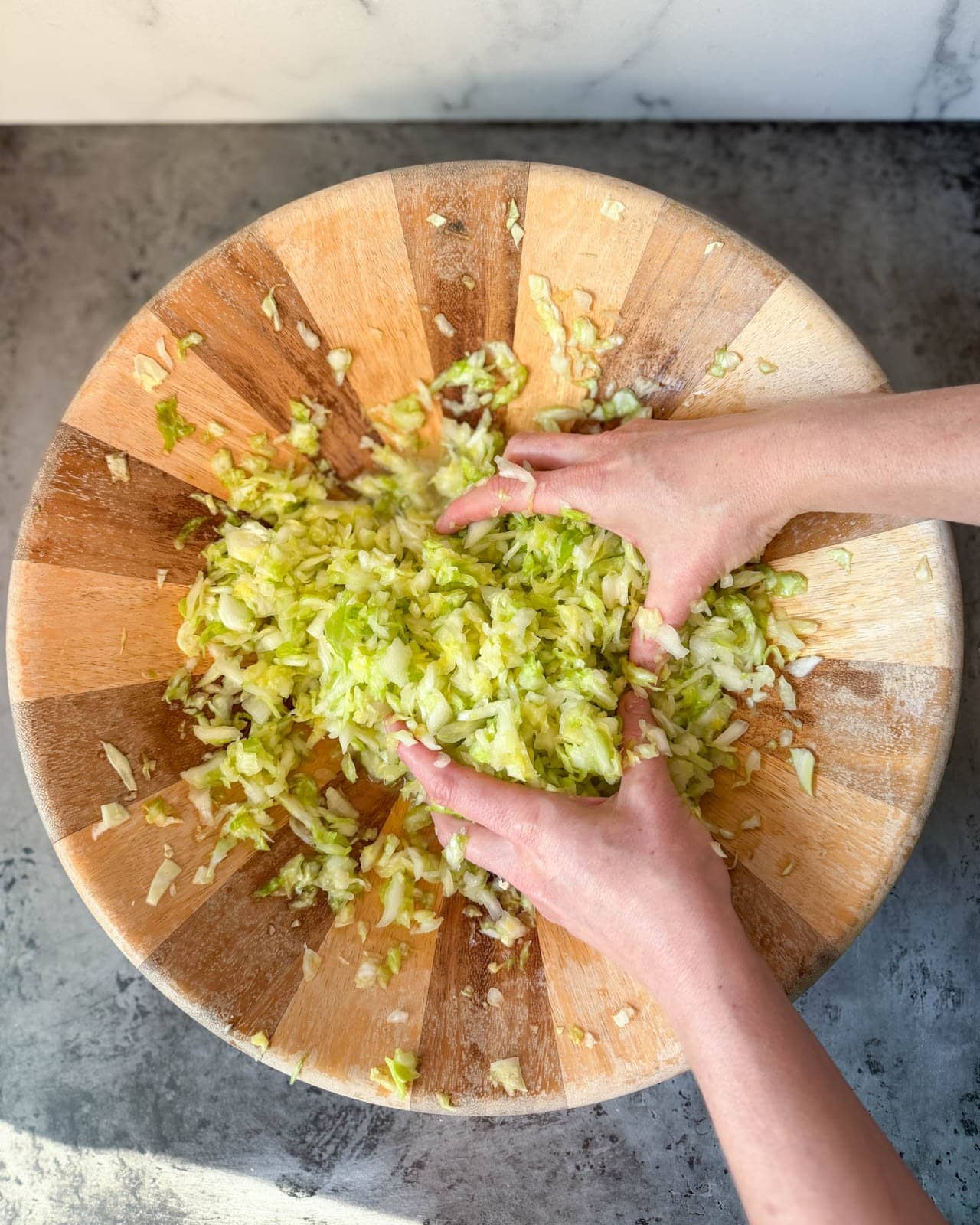 Two hands in a large wooden mixing bowl, massaging the salted cabbage that has now significantly softened, shrank in volume, and appears more wet and juicy than when raw