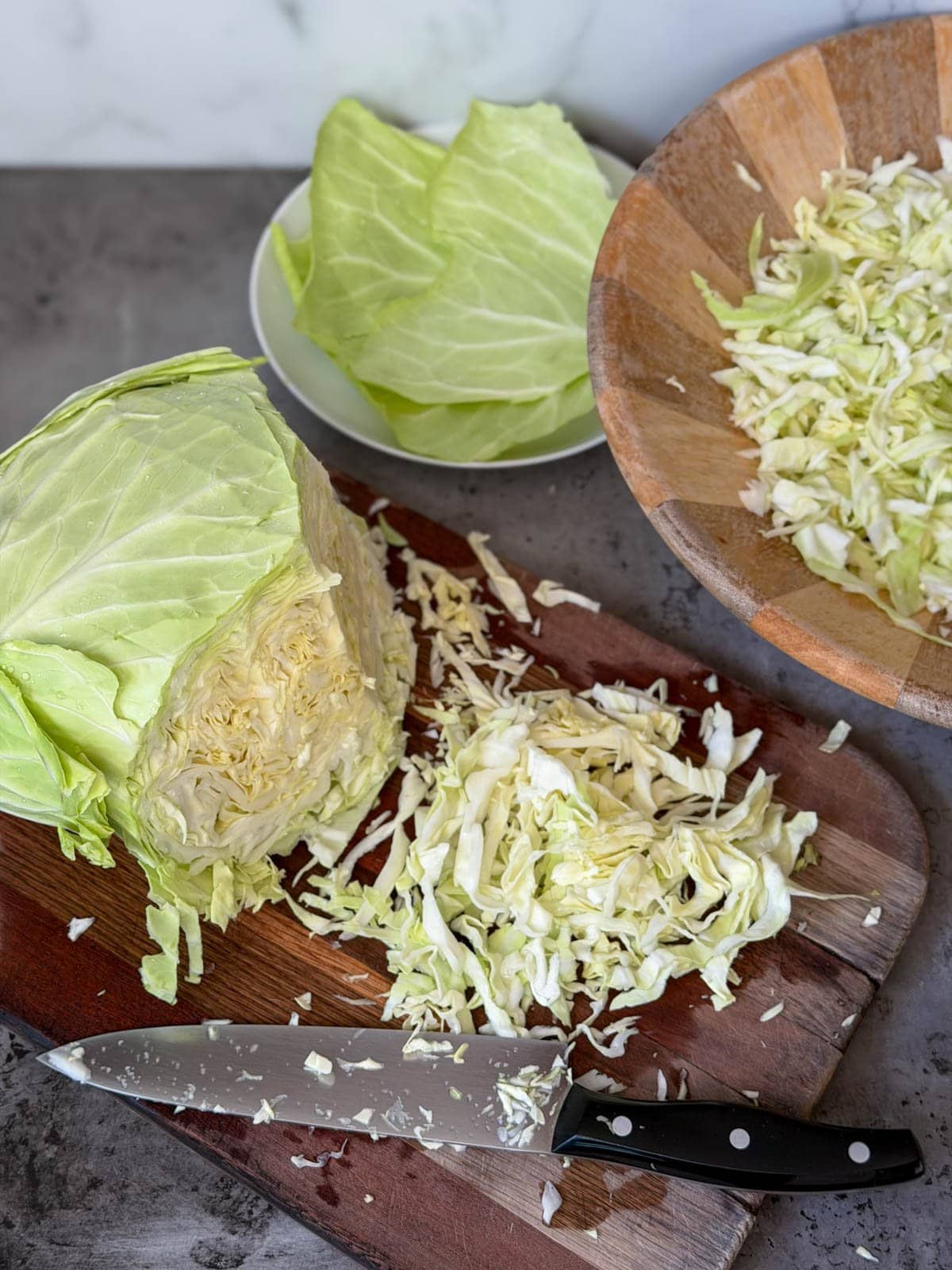 A head of cabbage and shredded pieces of cabbage sit on a wood cutting board. A large wood bowl is partially full of the shredded or cut cabbage.