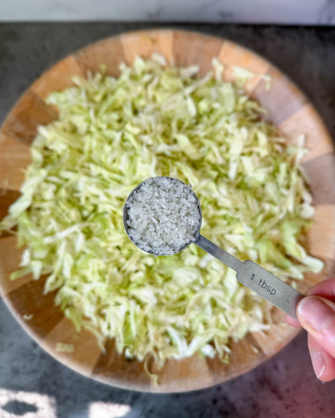 A close up a hand holding a tablespoon full of salt, hovering over the bowl of green shredded cabbage blurred below in the background