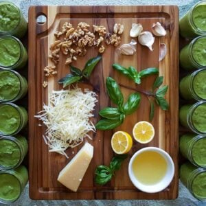 A wood cutting board with basil, walnuts, garlic, parmesan, and lemon juice arranged around the board. Small glass jars full of pesto line the outside of both sides of the cutting board.