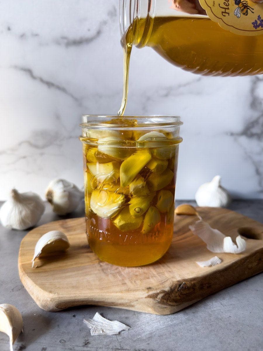 A pint mason jar is 2/3rds full of garlic cloves while honey is being poured into the jar, filling it almost all the way to the rim of the jar.