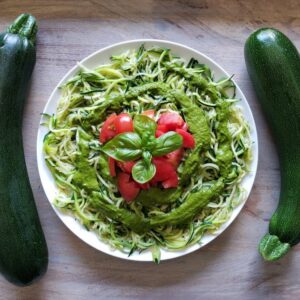 Two zucchini are aligned on each side of a large plate of pesto zucchini noodles or zoodles, chopped tomatoes and a sprig of basil are in the center of the bowl for garnish.