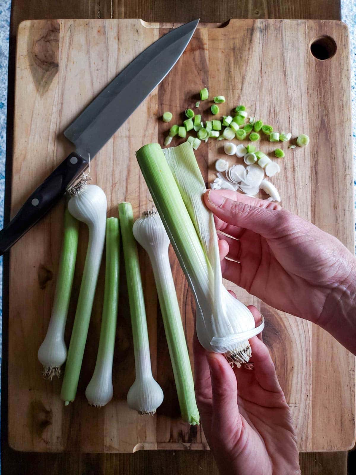 A hand is pulling away tough outer layers of the harvest bulbs and stems. A cutting board below contains more of the bulbs along with some that have been cut into pieces.