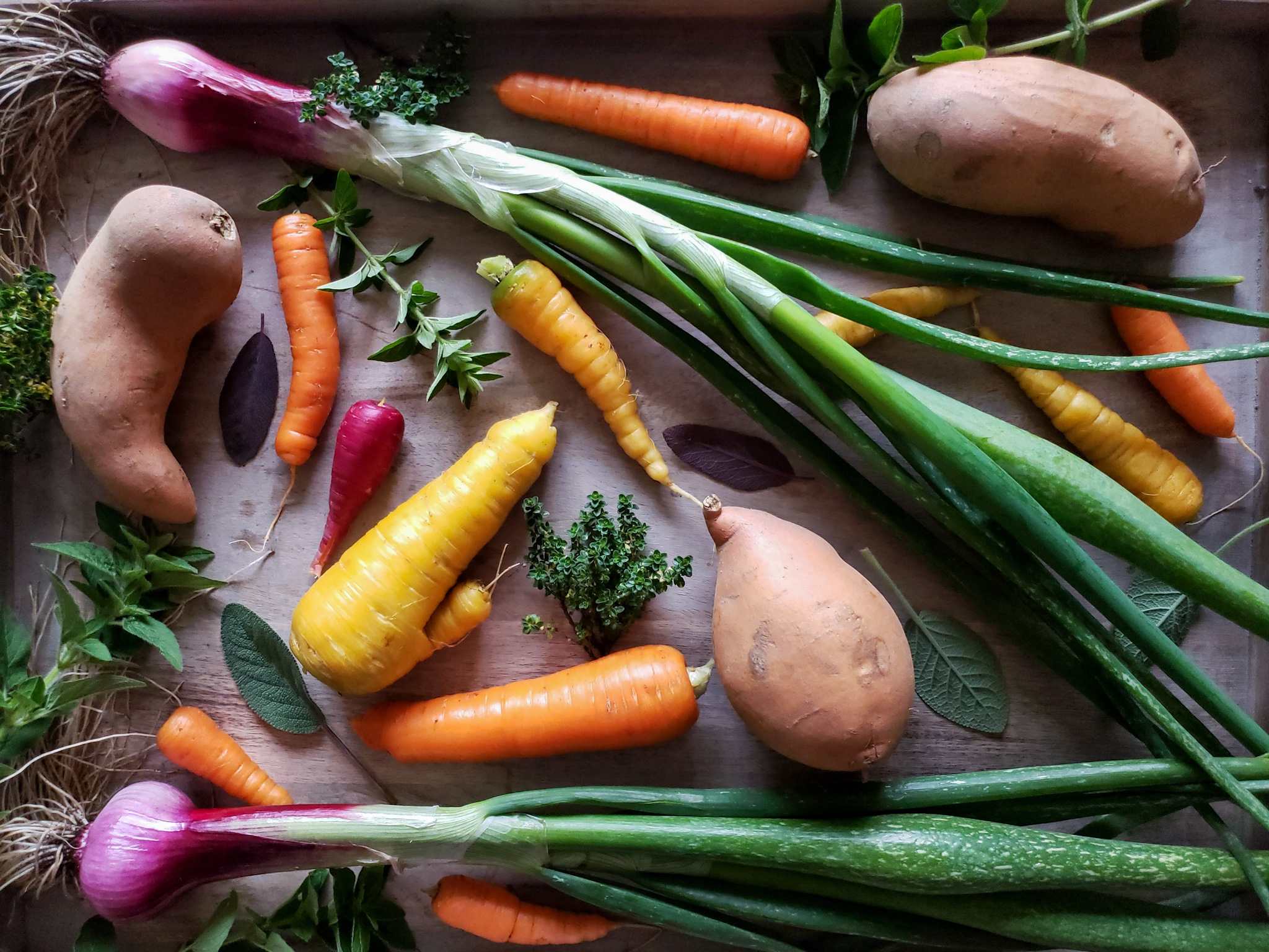 Looking down on some of the fresh ingredients used in this recipe, artfully laid out on a cutting board in moody lighting. There are several sweet potatoes, carrots, red onions with their greens still attached, and a variety of fresh herbs on display.