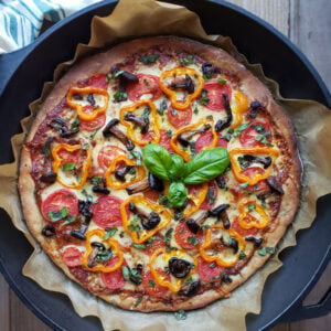 A close up of a just-baked sourdough pizza still in the cast iron pan. The pizza looks perfectly baked with a golden-brown crust, topped with sliced tomatoes, cheese, rings of orange bell peppers, mushrooms, and a sprig of fresh basil in the center