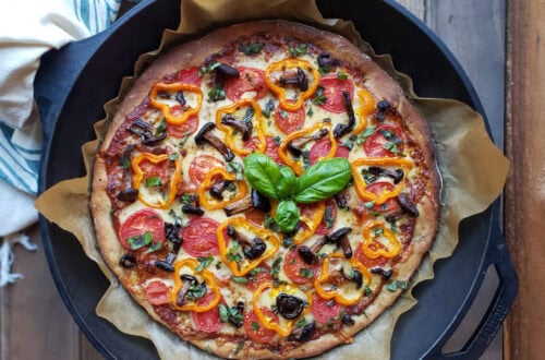 A close up of a just-baked sourdough pizza still in the cast iron pan. The pizza looks perfectly baked with a golden-brown crust, topped with sliced tomatoes, cheese, rings of orange bell peppers, mushrooms, and a sprig of fresh basil in the center