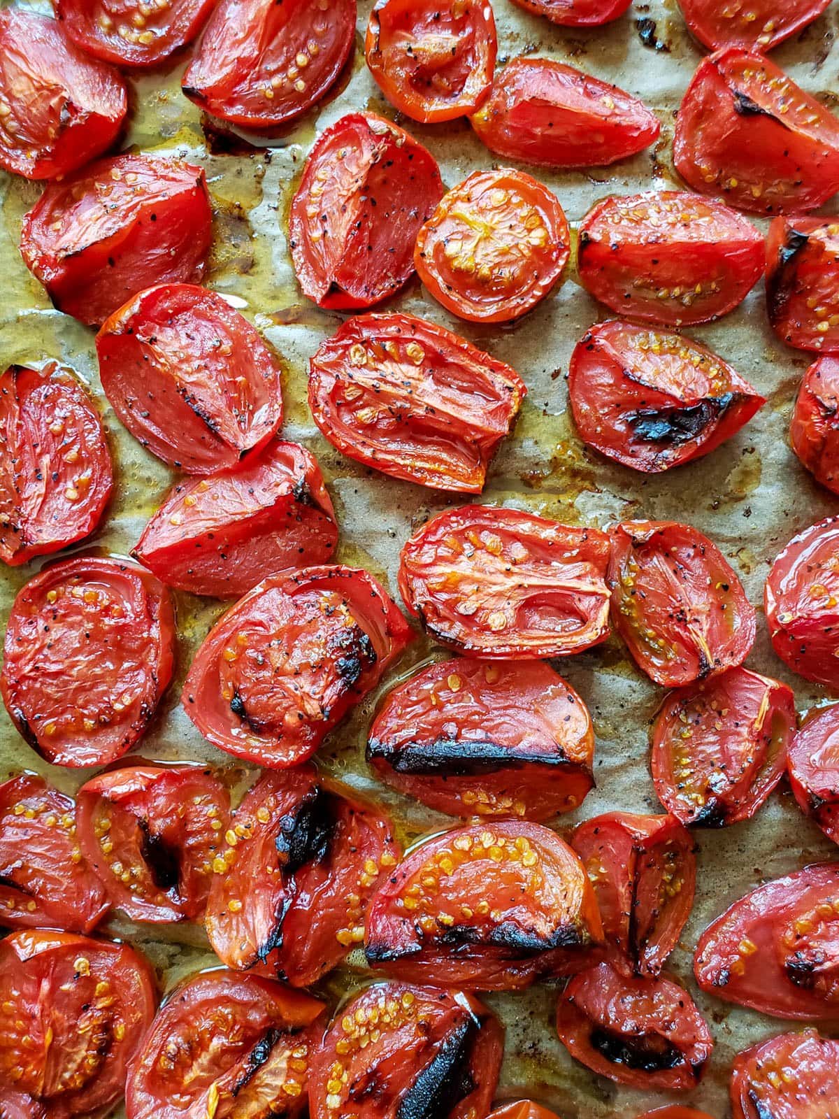 A close up of a baking sheet full of red tomatoes cut into quarters after roasting, with browned spots and slightly shriveled and juicy