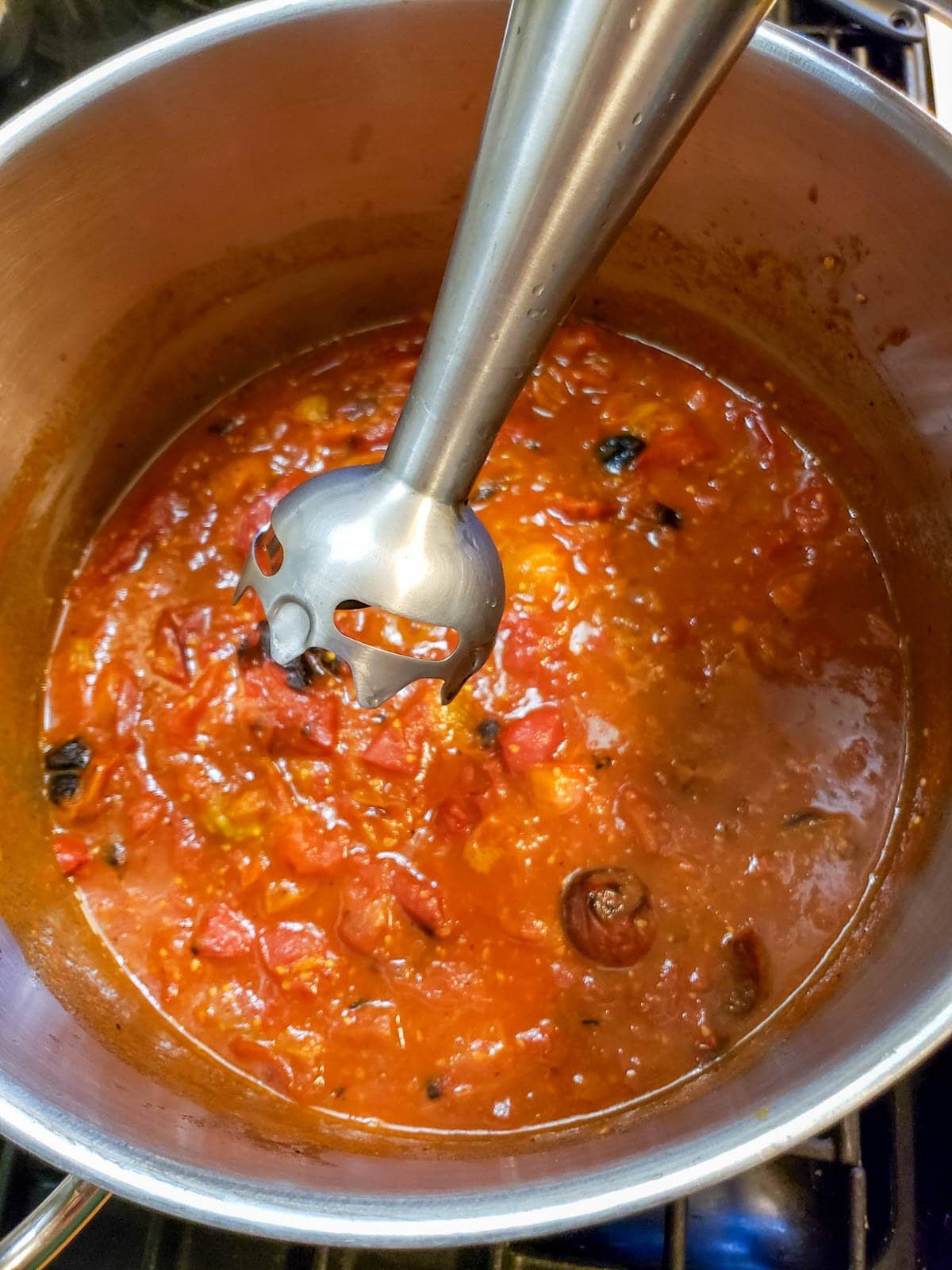 An immersion blender is held above a pot of stewed and roasted tomatoes.
